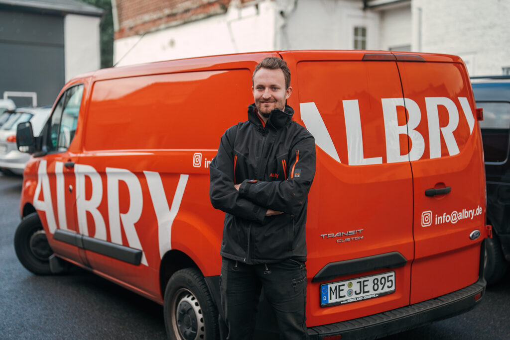 Timo Albry steht vor dem orangefarbenen Firmenfahrzeug
